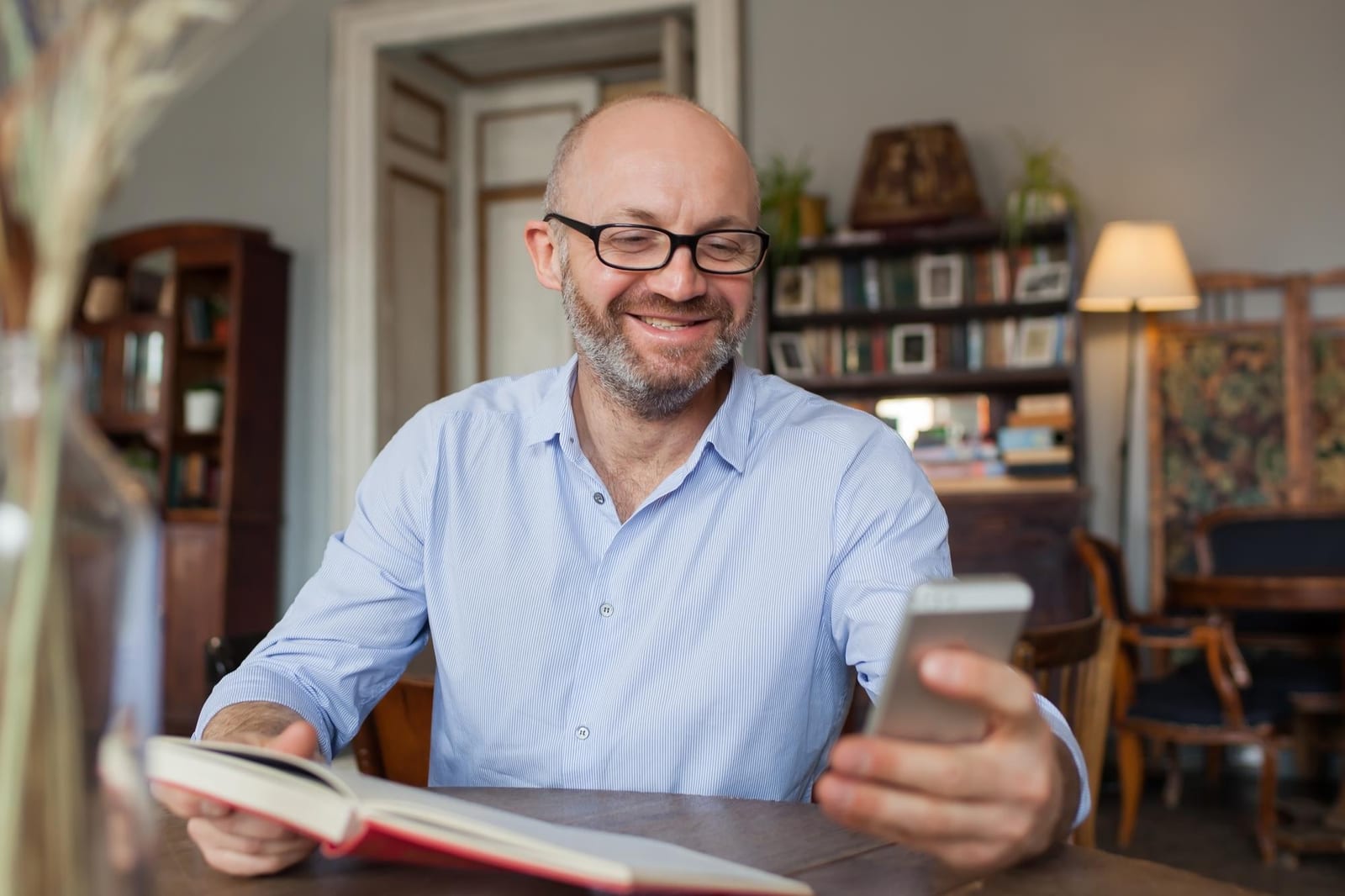 Homem lendo um livro de capa vermelha e consultando o celular em uma sala com estante de livros.