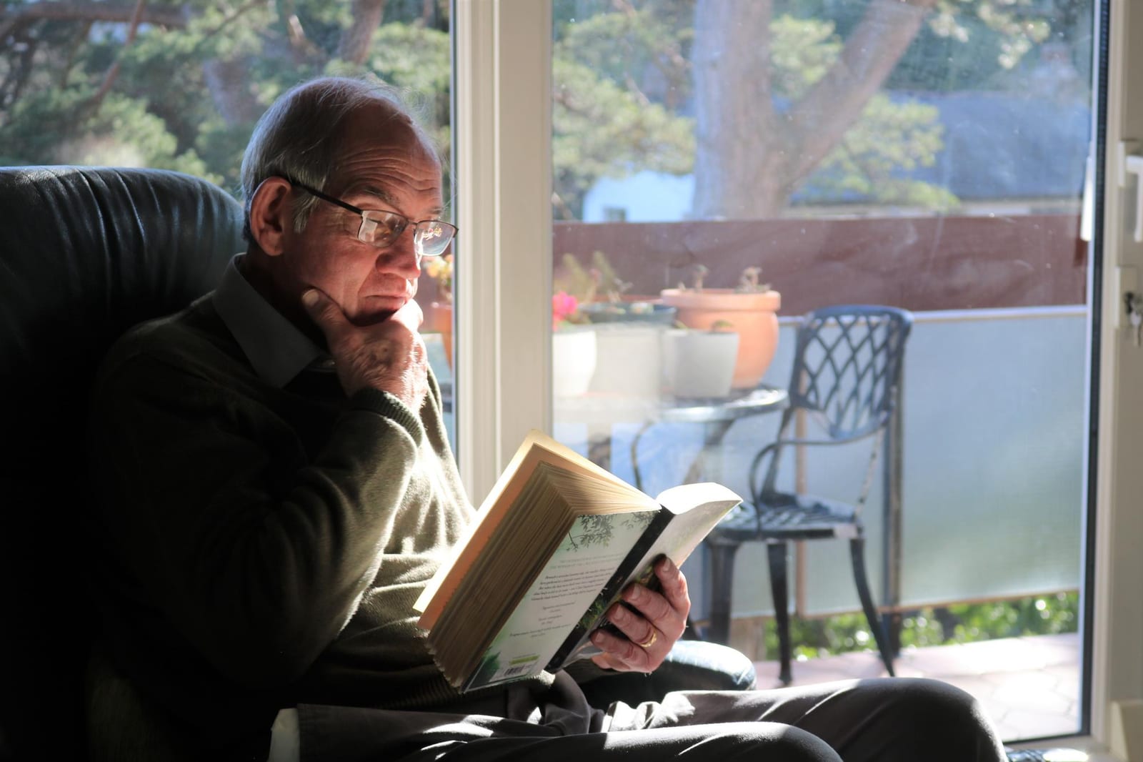Homem lendo um livro em pé, junto a uma janela ampla com vista para um terraço.