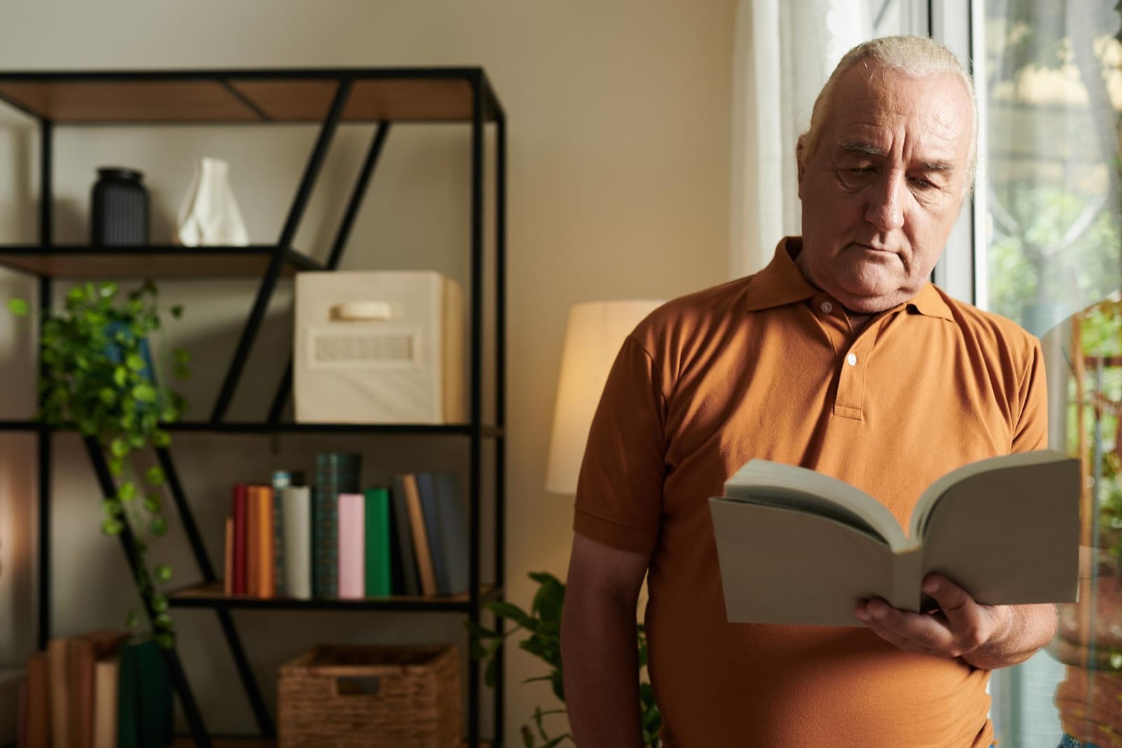 Homem em pé lendo um livro perto de uma estante com plantas ao fundo.