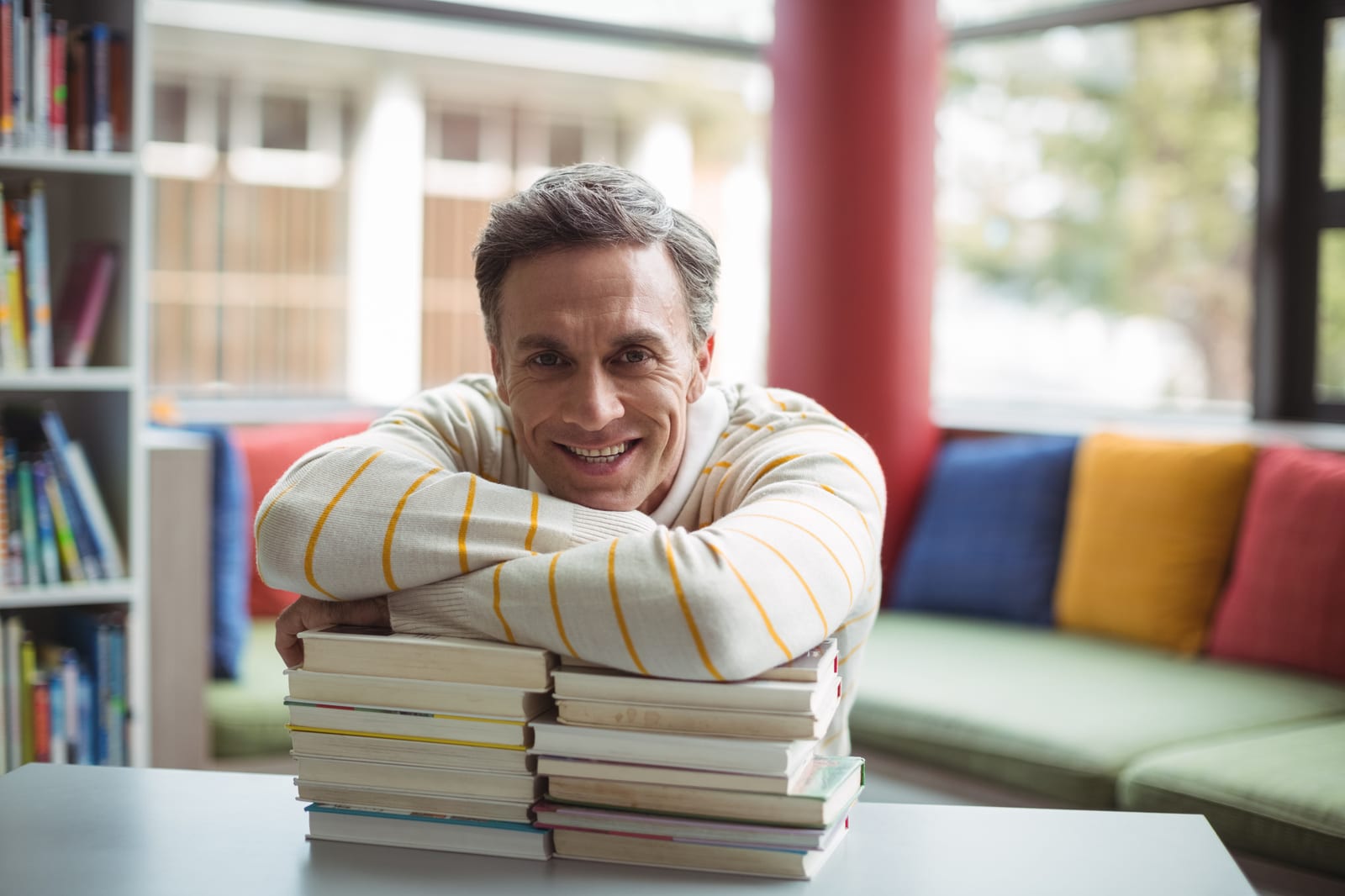 Homem sorridente apoiado sobre uma pilha de livros, em um espaço de leitura com sofá colorido ao fundo.